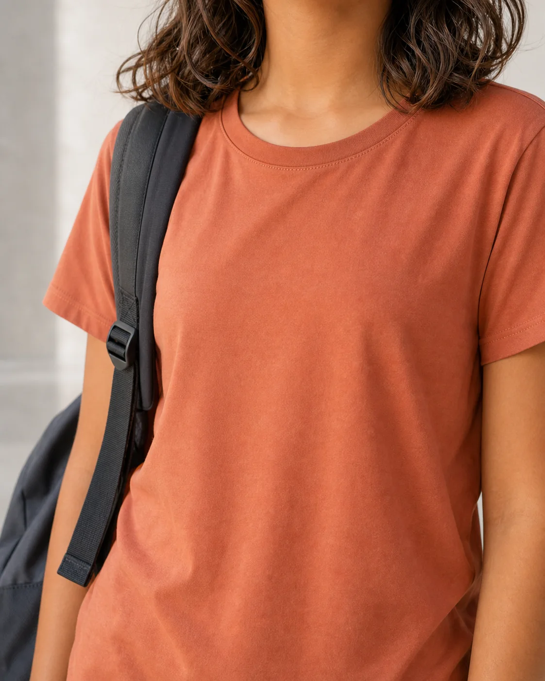 Girl from the collarbone down wearing a plain terracotta t-shirt with a backpack strap over one shoulder